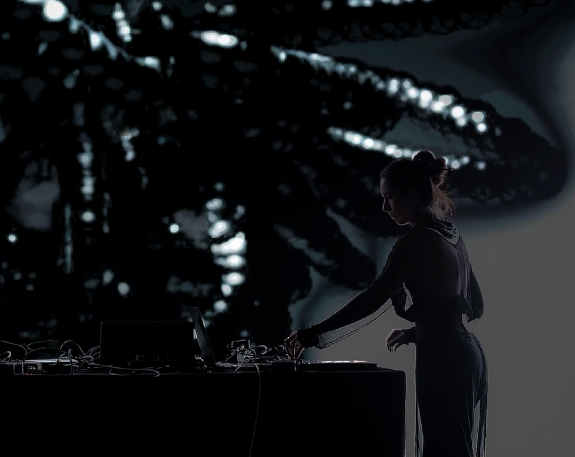 Ninon in profile at a desk of laptops and controllers, backlit by a large generative projection
