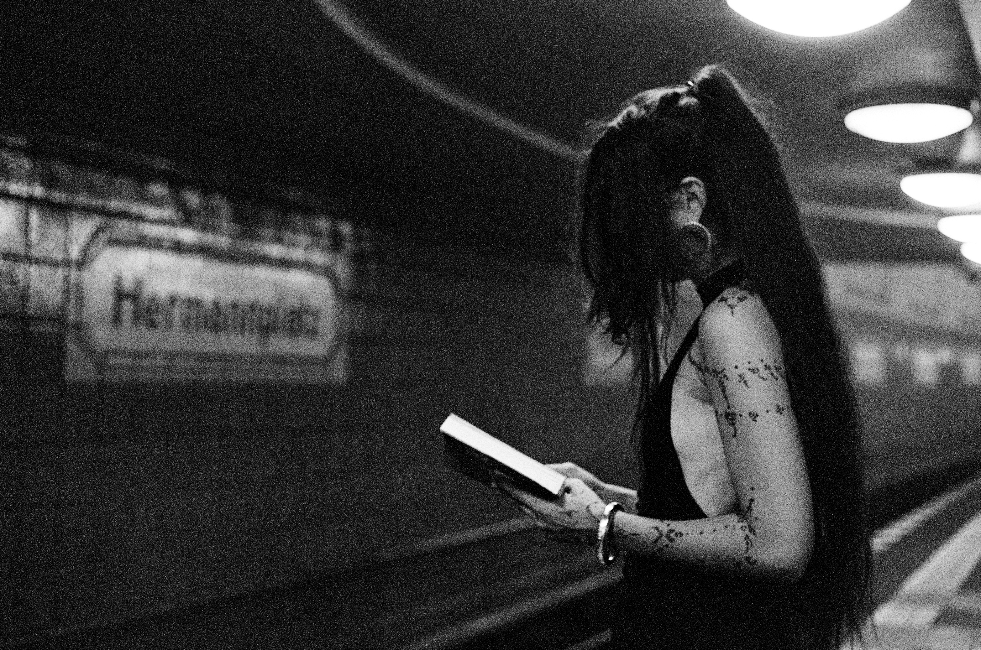 Black and white photograph: Ninon reading a book on a Berlin U-Bahn platform, the station sign for Hermannplatz blurred behind her