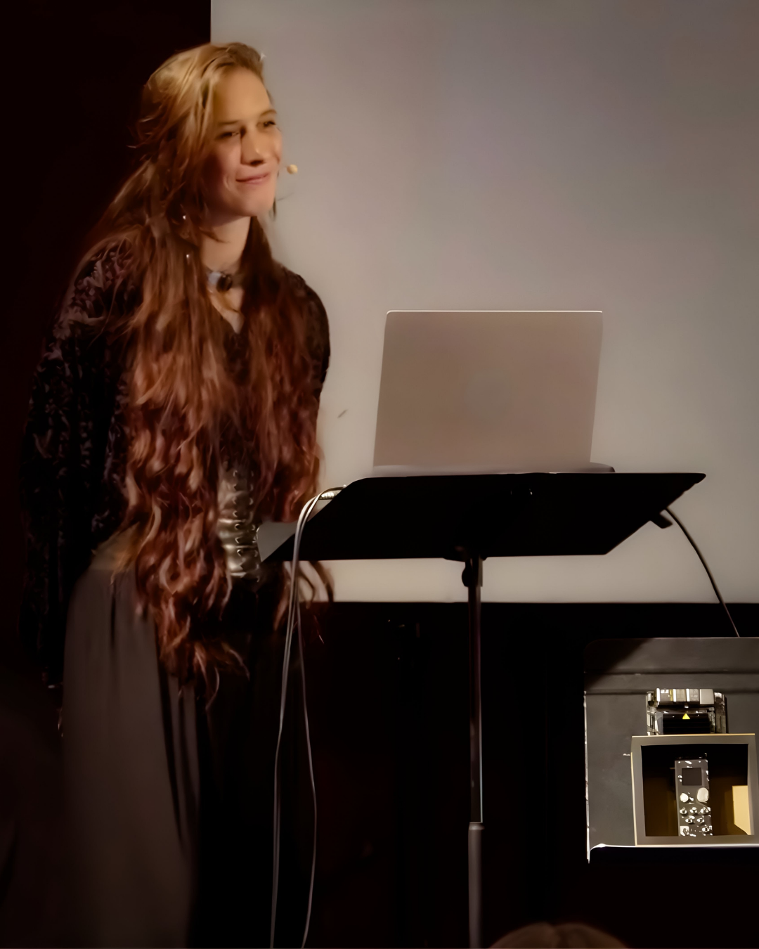Ninon presenting at the thesis defence, wearing a headset microphone beside a music stand holding a laptop, with the Neurorack module visible on a table to the right