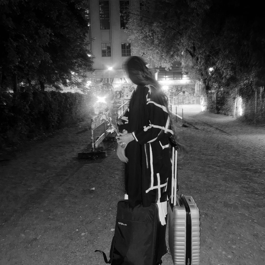 Black and white photograph: Ninon seen from behind on the path outside Berghain at night, in a long patterned coat, holding flowers, a modular synthesiser case and a suitcase standing beside her, the lit facade of the building behind the trees