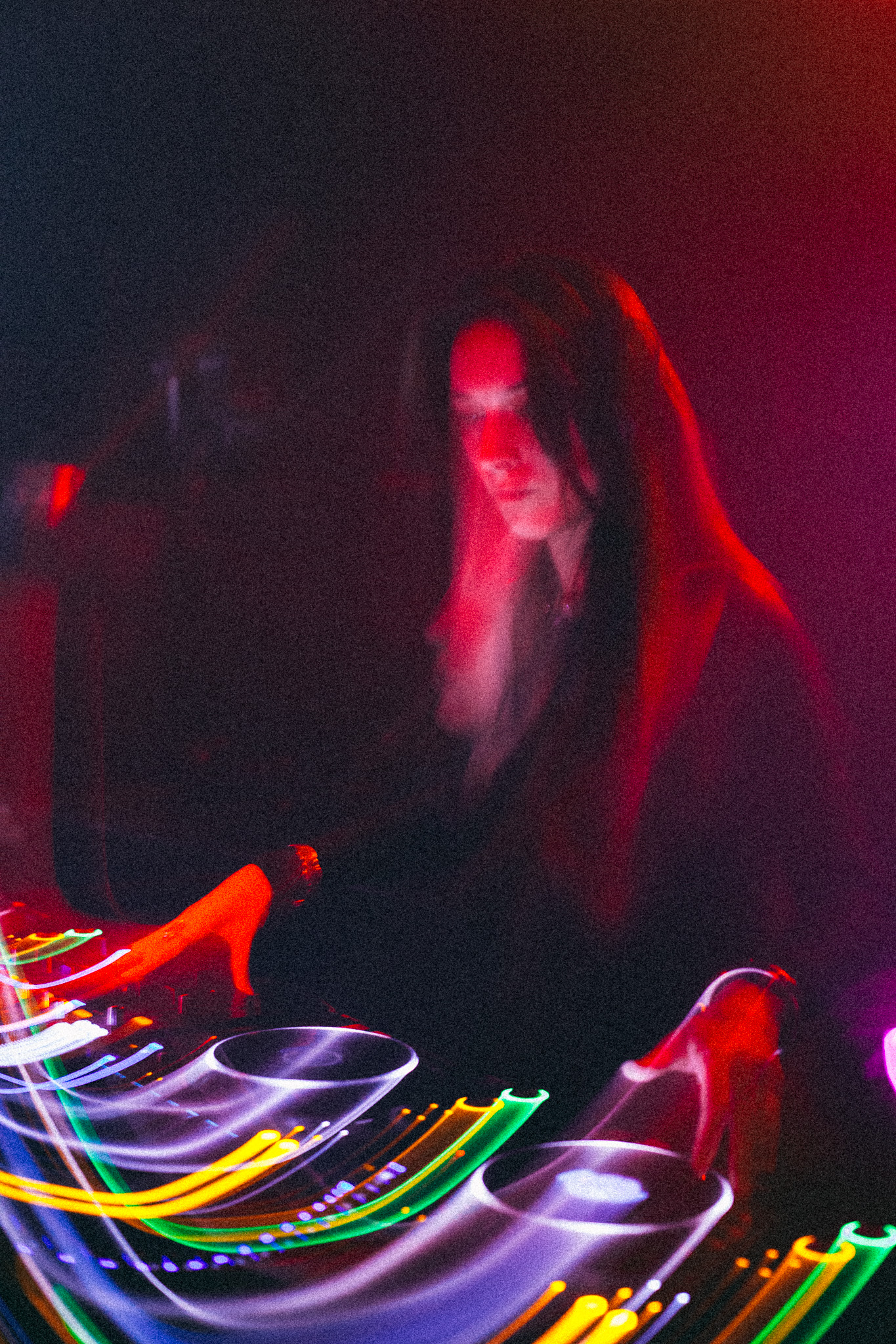 Ninon behind the decks in red and magenta light, the frame streaked with the motion of the mix
