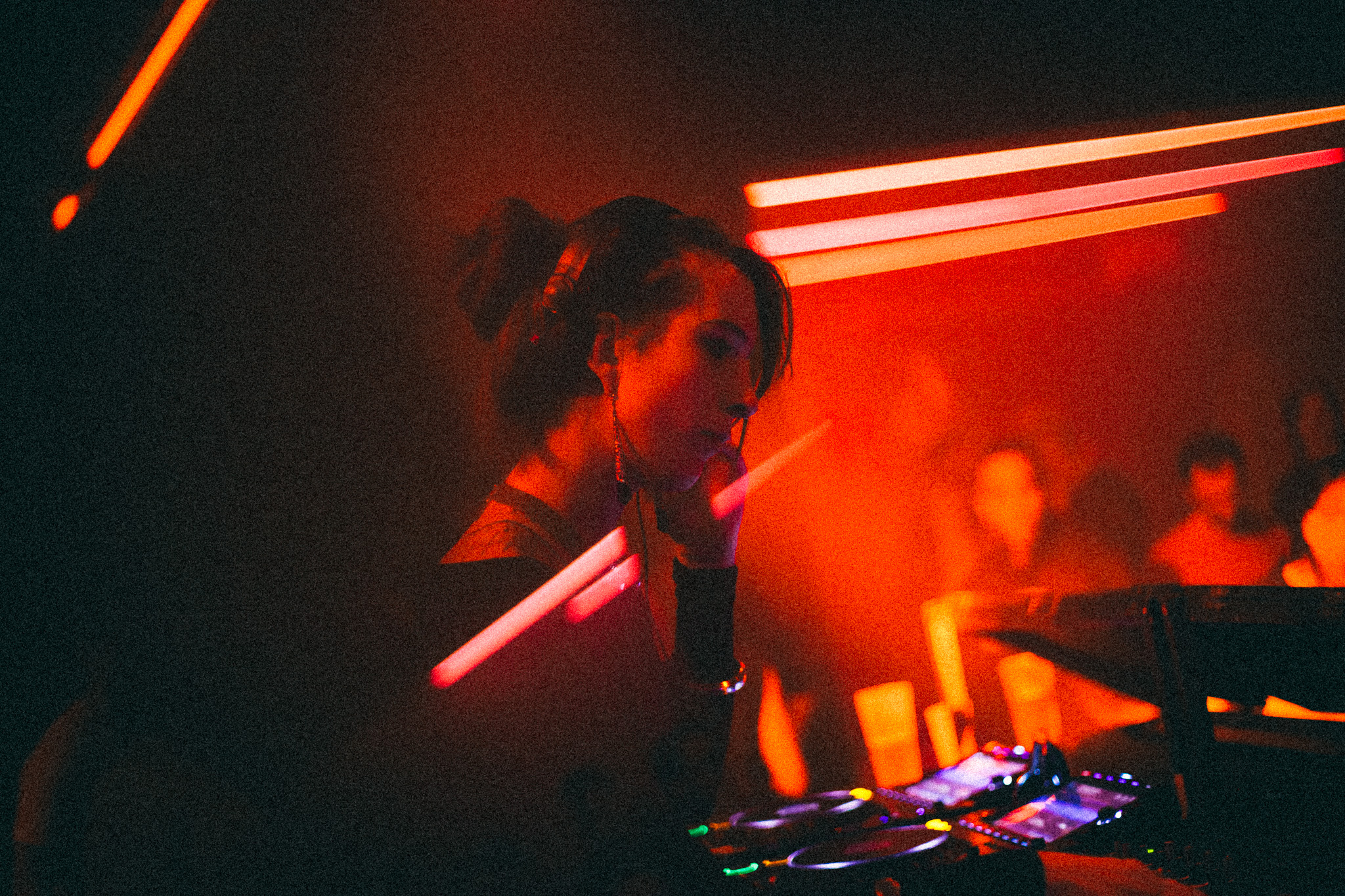 Ninon close to the decks, horizontal red neon bars behind, the crowd beyond