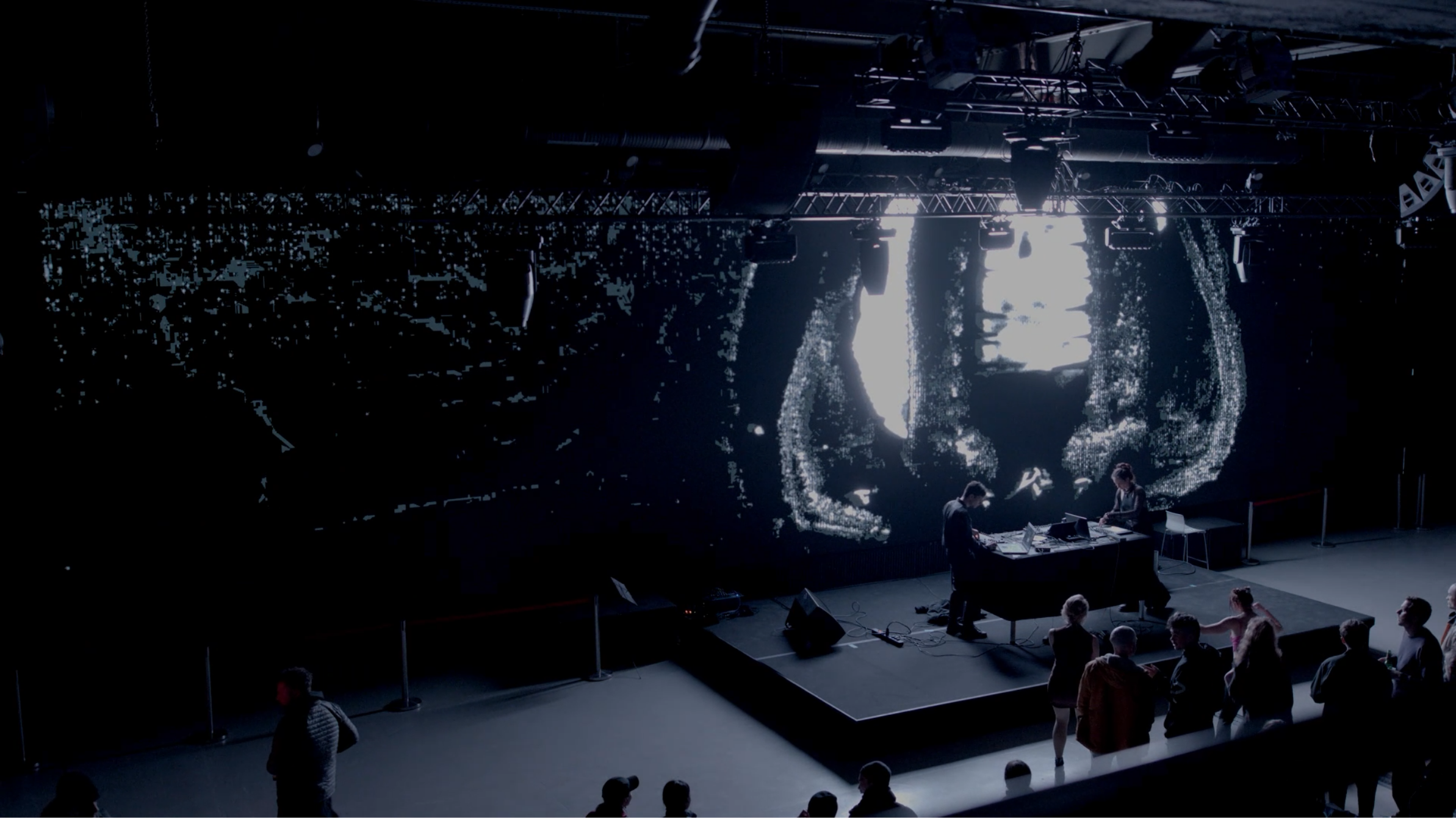The room at Alte Münze from the balcony: the projection has shifted to a dense vertical structure, the audience gathered close to the stage