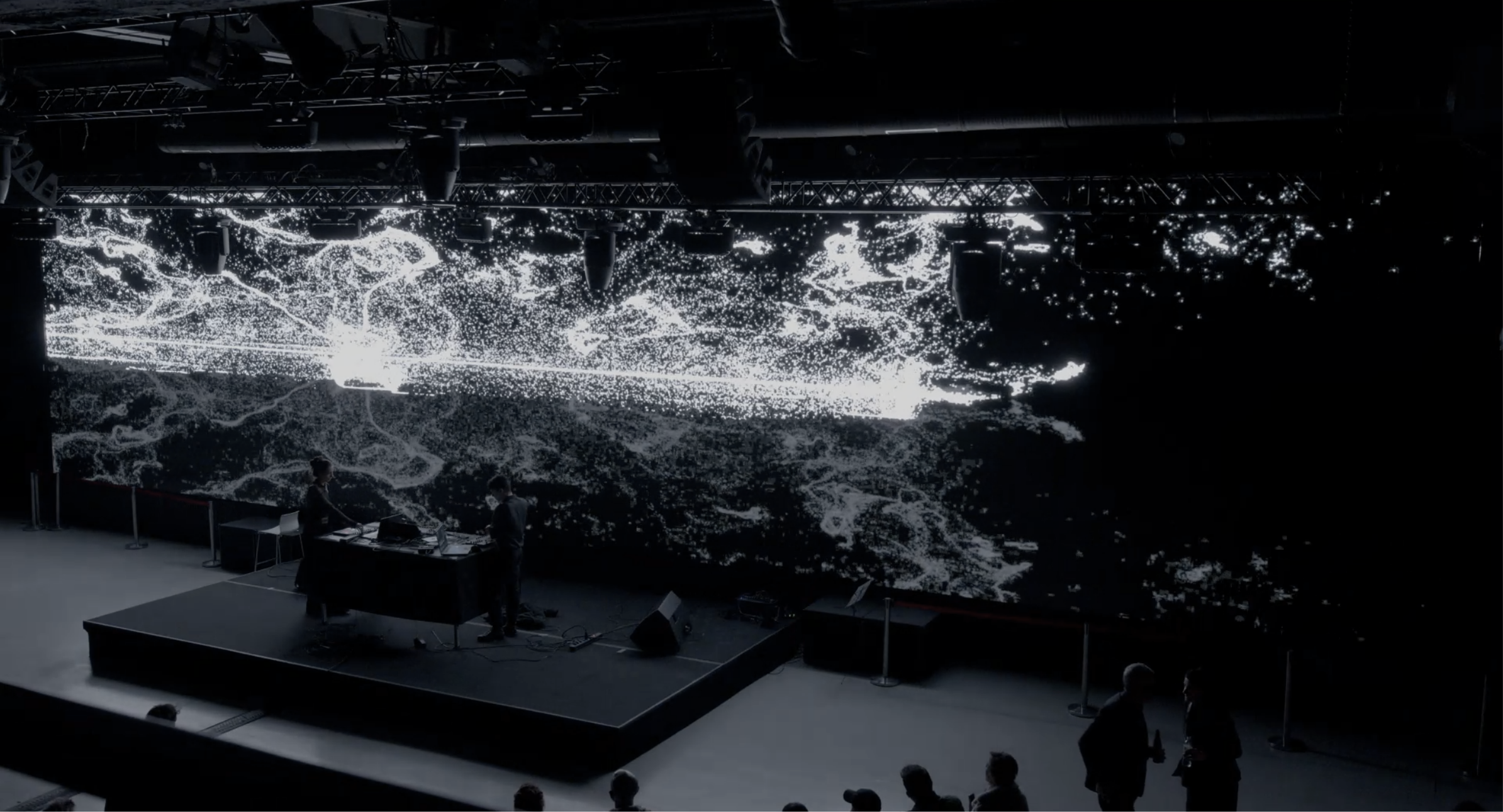 Wide view of Alte Münze: two performers on a low stage in front of a wall-sized projection of a bright particle field, audience in the foreground