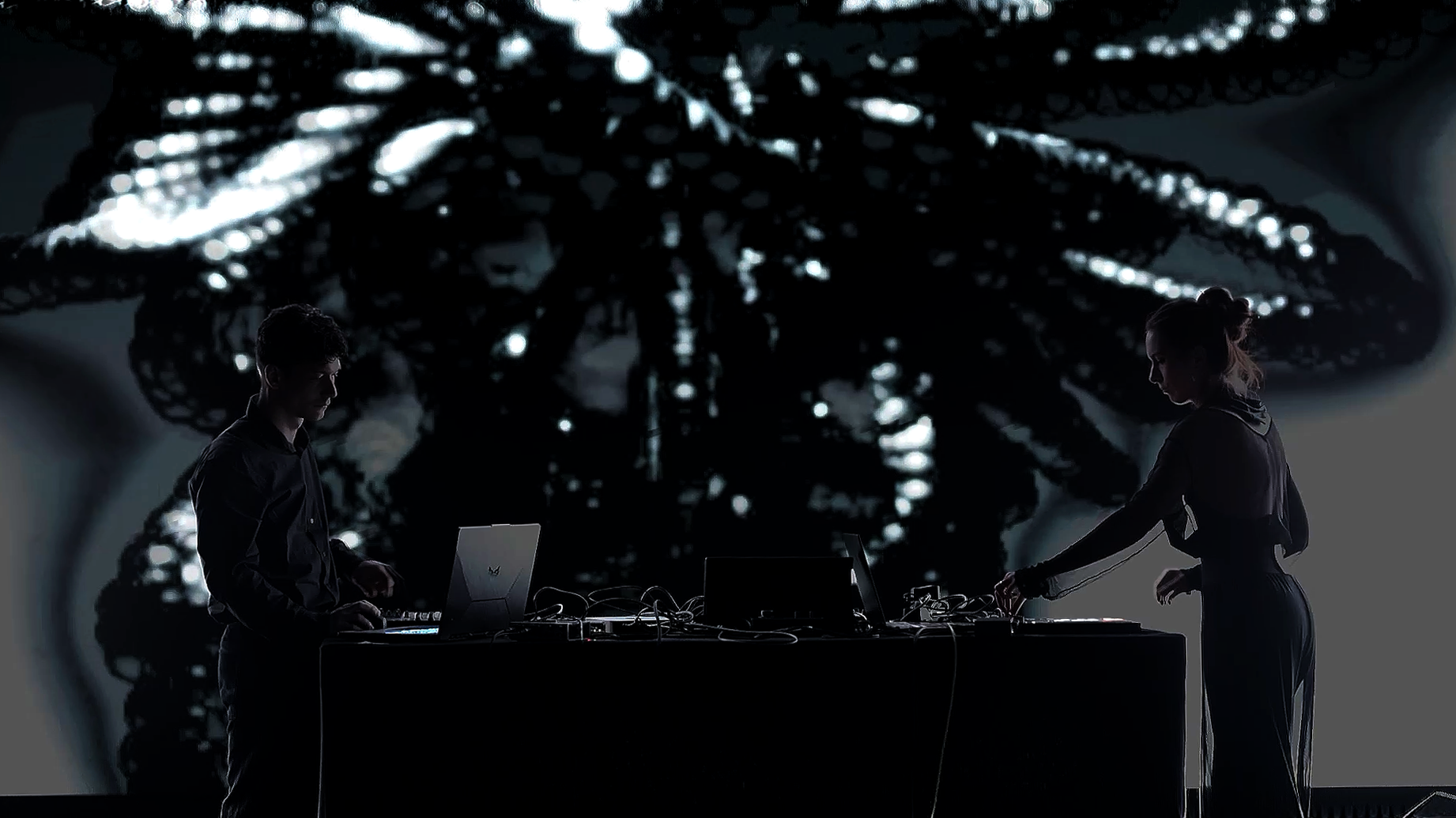 Ninon and Victor Calvi in silhouette behind a desk of laptops and controllers, backlit by a large generative projection