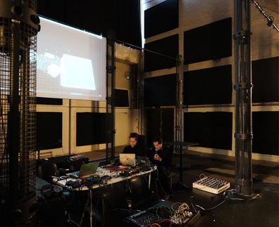 Ninon and Jun Suzuki working at a desk of laptops and modular gear inside the MONOM 4DSOUND room, a projection lit behind them